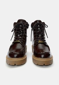 Bruine leren enkelbotten met een glanzende afwerking, bruine rubberen zool, ronde neus en traditionele vetersluiting. Gouden accent op de tong.