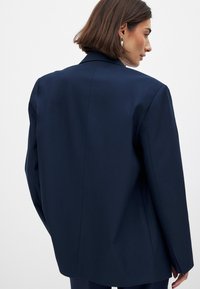 Blazer ajusté bleu marine avec une silhouette structurée, présentant un col à revers et un tissu lisse ; peu de détails et un design arrière épuré.