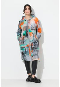 Kleurvolle, capuchonjas met abstracte patronen in grijs, oranje en groen. Heeft een ritssluiting en zijzakken. Gedragen over zwarte leggings en laarzen.