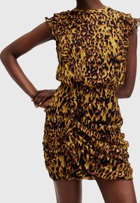 Kvinna iklädd en rynkad mini-klänning med leopardmönster, volang ärmar och metalliska detaljer, poserande med en hand på höfterna.