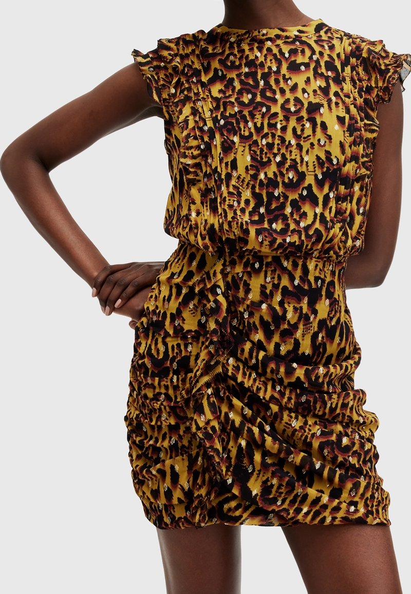 Kvinna iklädd en rynkad mini-klänning med leopardmönster, volang ärmar och metalliska detaljer, poserande med en hand på höfterna.