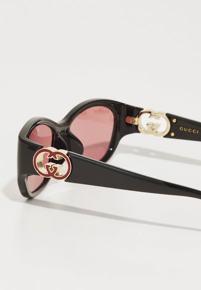 Slnečné okuliare Gucci s čiernym plastovým rámom, ružovými šošovkami a zlatými detailmi, pričom na spánkoch sú umiestnené akcenty prepojeného loga G.
