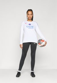 T-shirt de sport à manches longues blanche avec un logo bleu et rouge, associé à un legging noir avec des accents texturés, et des chaussures de sport noires tenant un ballon de football.