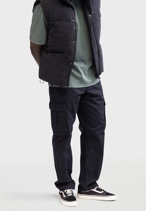Homme portant un pantalon cargo noir, un gilet en velours côtelé noir, un t-shirt vert et des baskets noires et blanches, debout les mains dans le dos.
