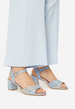 Niebieskie sandały na klockowym obcasie z metalicznymi srebrnymi akcentami, wyposażone w krzyżujące się paski i zapięcia wokół kostki. Gładka tekstura i nowoczesny design.