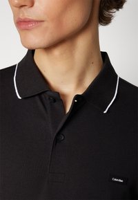 Čierna polo košeľa z textúrovaného materiálu s bielym lemovaním goliera, so zapínaním na dve gombíky a malou nášivkou loga Calvin Klein.