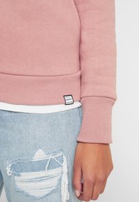 Mano derecha relajada al lado, vistiendo una camiseta de manga larga rosa y jeans desgastados de color azul claro con un parche deshilachado en el muslo.