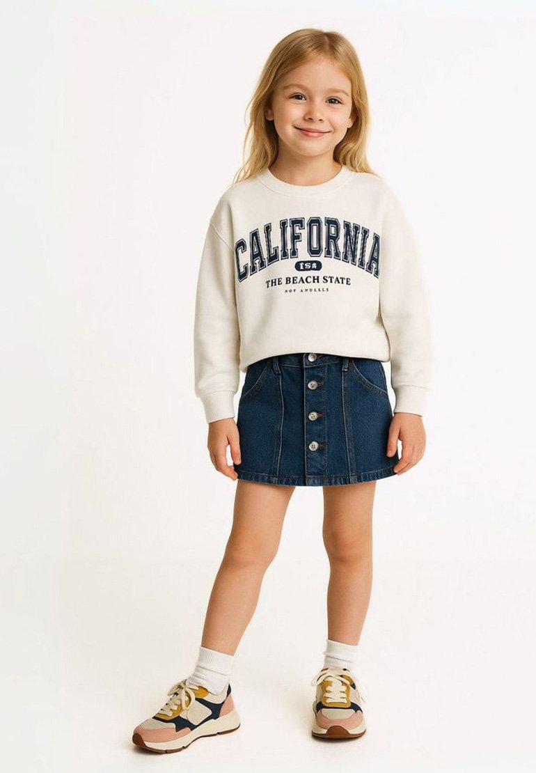 Felpa bianca con stampa "CALIFORNIA", gonna A-line in denim blu con bottoni frontali e sneakers beige con accenti blu navy e senape.