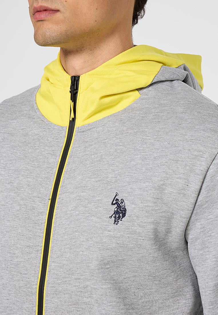 Hoodie zippé gris avec capuche jaune. Présente un logo noir brodé sur la poitrine. Fabriqué en tissu texturé avec des détails de fermeture éclair contrastants.