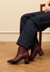 Bordeaux lederen enkellaarsjes met een spitse neus en een blokhak, gecombineerd met losse donkere denim jeans, op een houten vloer.