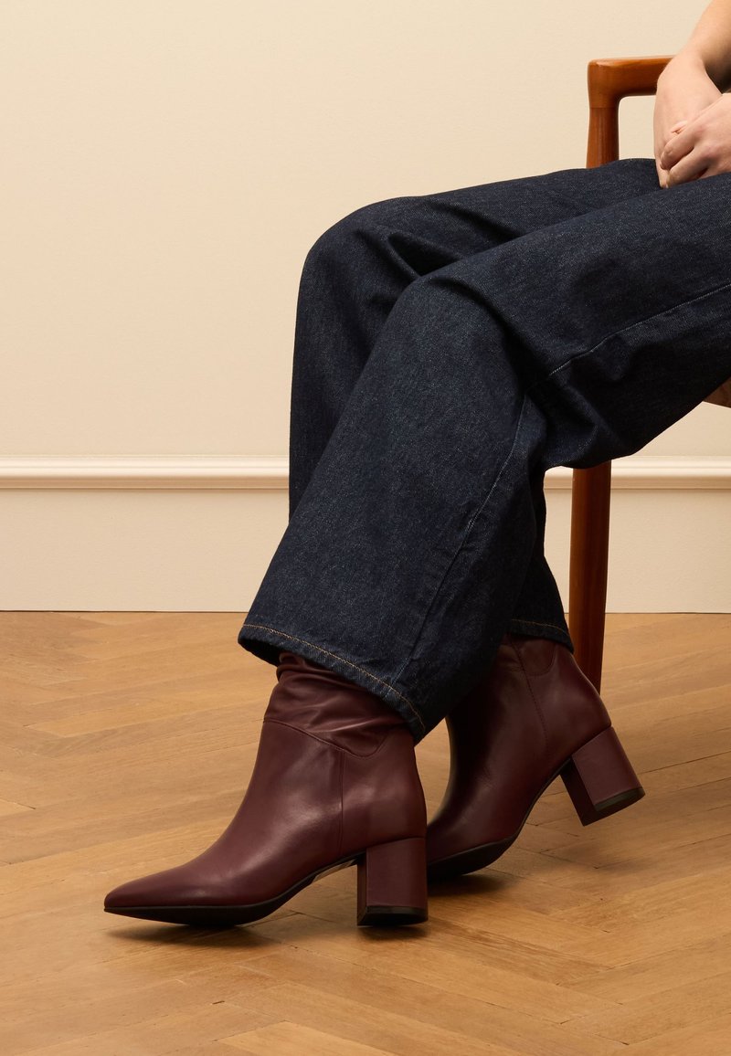 Bordeaux lederen enkellaarsjes met een spitse neus en een blokhak, gecombineerd met losse donkere denim jeans, op een houten vloer.