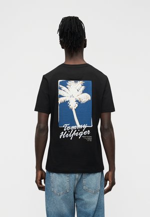 Uomo che indossa una T-shirt nera con una grande palma bianca e la scritta "Tommy Hilfiger" sul retro, abbinata a jeans azzurri.