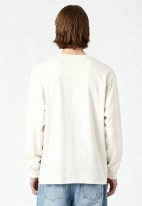 Homme aux cheveux bruns mi-longs portant une chemise blanche à manches longues unie et un jean bleu clair, vu de dos sur un fond blanc.