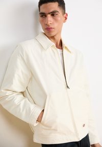 Cream corduroy jacket with a classic collar, front zipper, and side pockets. Features subtle stitching and a small logo detail at the hem.