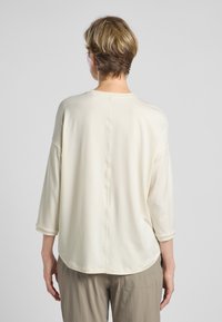 Cremefarbenes Langarmshirt mit lockerer Passform, abfallenden Schultern und abgerundetem Saum. Glattes Material mit subtilen Nähdetails auf der Rückseite.