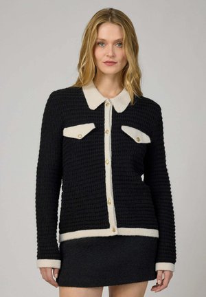 Femme blonde portant un cardigan noir texturé avec col, poignets et poches blancs, ainsi que des boutons dorés, assorti d'une jupe noire.
