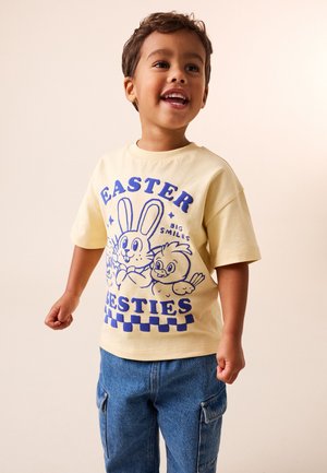 Smilende ung dreng iført en blegegul påske-tema t-shirt med blå tegneserie-kanin og kylling samt blå jeans.