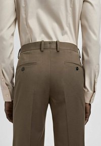 Braune, maßgeschneiderte Hose mit glatter Textur, zwei Gesäßtaschen, Knopfdetails und einem eleganten Bund, kombiniert mit einem hellbeigen Hemd.