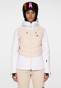 Osoba w beżowej i białej kurtce narciarskiej oraz spodniach, czarnym kasku narciarskim i lustrzanych goglach narciarskich, stojąca przodem na jednolitym tle.