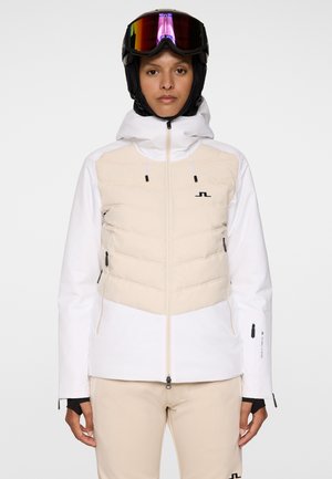 Person i beige og hvid skijakke og -bukser, sort skihjelm og reflekslignende skibriller, stående fremadrettet mod en ensfarvet baggrund.