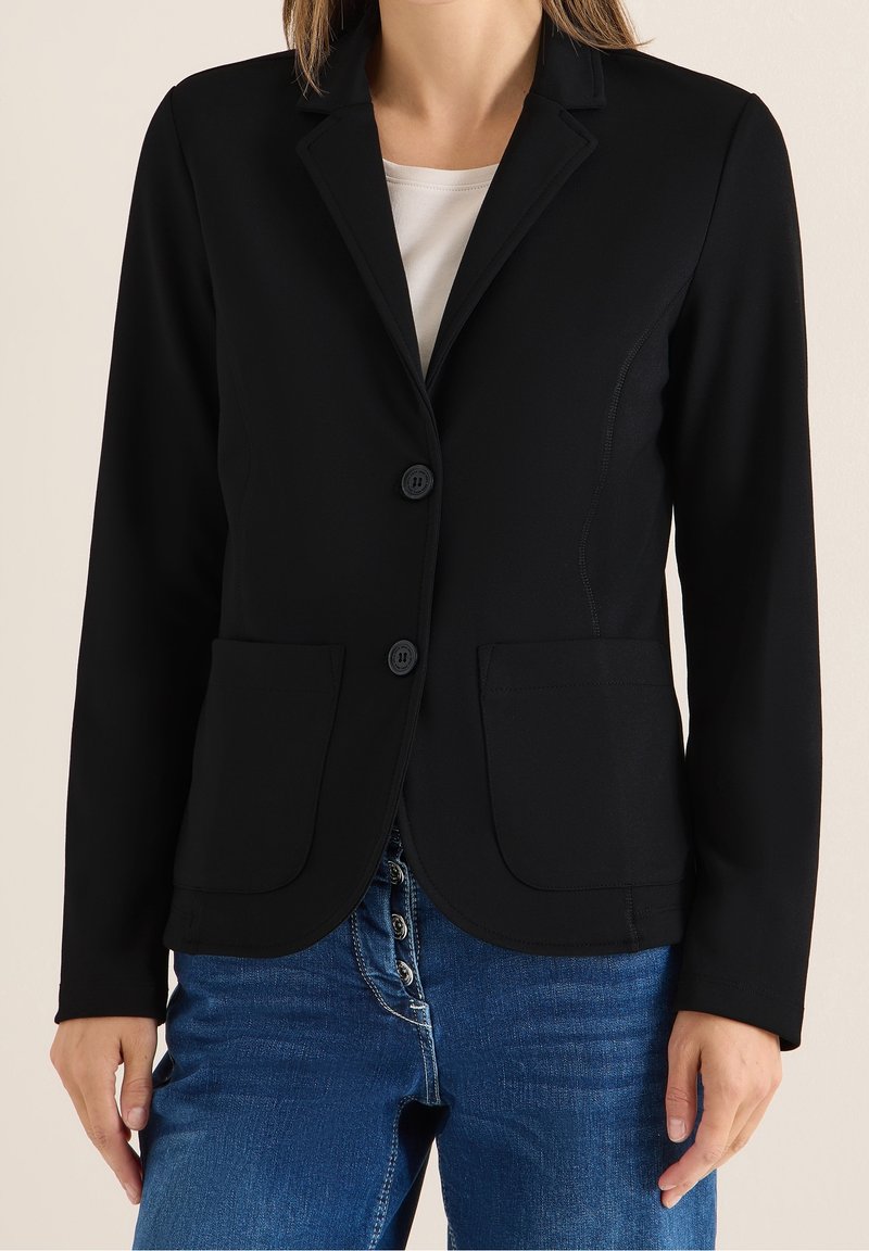 Femme portant un blazer noir boutonné sur une chemise blanche et un jean bleu, mains détendues le long du corps.