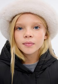 Čierna puffa bunda s vysokým golierom, doplnená o mäkkú bielu kožušinovú čiapku. Svetlo blond vlasy a bledá pleť zvýrazňujú celkový vzhľad.