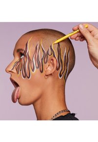 Ett calvhuvud med färgglad ansiktsmålning i flammmönster i brunt, gult och vitt. En hand håller en gul penna nära den målade hårlinjen.