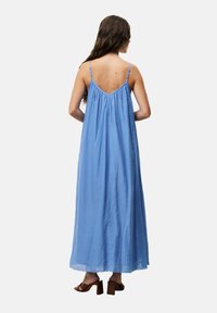Lichtblauwe maxi-jurk met dunne verstelbare bandjes, geraffineerde stof aan de achterkant, vloeiende textuur en een lichte glans. Bruine blokhakken sandalen.