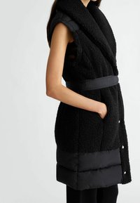Czarna kamizelka bez rękawów z miękką, puszystą teksturą zewnętrzną, eleganckim czarnym dołem pikowanym, talią ściągniętą i bocznymi kieszeniami.