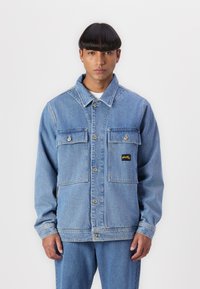 Blue denim jacket with two chest pockets, a button closure, and a small yellow logo patch on the left pocket. Features a collared design.