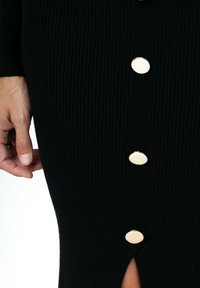 Robe noire côtelée avec quatre grands boutons ronds sur le devant, et une fente en bas. Texture douce et design ajusté.