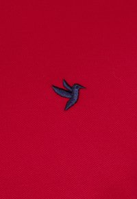 Un tissu textile rouge présente un petit logo de colibri brodé en bleu marine, positionné sur le côté gauche. La texture est légèrement relevée.