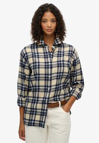 Chemise en flanelle à carreaux bleu marine et beige avec des manches longues, un col boutonné et une poche ; portée avec un pantalon beige et une ceinture marron.