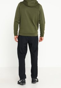 Sudadera con capucha de color verde oscuro con una textura suave, combinada con pantalones deportivos negros. Presenta puños elásticos y un corte relajado. Zapatillas negras visibles.