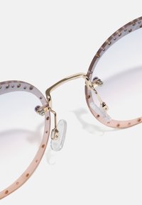 Occhiali da sole rotondi con lenti sfumate, montatura trasparente di un rosa chiaro, dettagli in metallo dorato e pois decorativi lungo il bordo superiore.