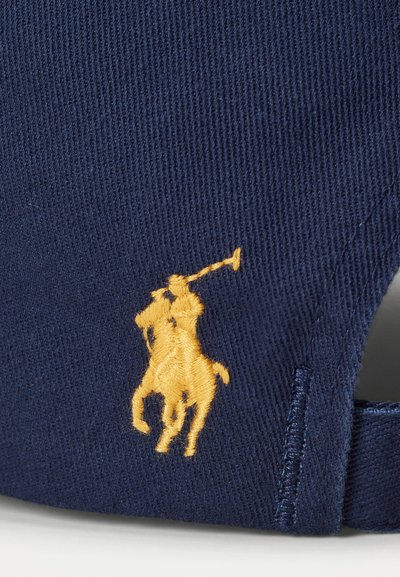 Tamsiai mėlyna medvilninė kepurė, priekyje su geltona siuvinėta polo žaidėjo logotipu. Reguliuojama galinė dirželio su siuvinėjimo detalėmis.