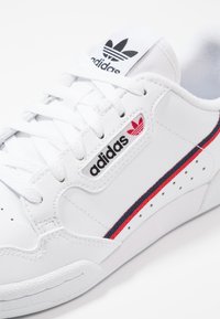 Sneaker sintetiche bianche con logo Adidas nero e rosso. Accenti a righe distintive e suola in gomma testurizzata con perforazioni.