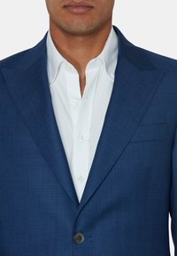 Niebieski żakiet uszyty z teksturowanej tkaniny, z klapami, pojedynczym zapięciem na guzik i kieszenią na piersi, noszony na białej koszuli.