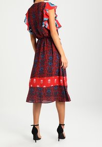 Frau trägt ein rot-blau gemustertes Midikleid mit schwingenden Ärmeln, steht in schwarzen High-Heel-Ankle-Strap-Schuhen und schaut weg.