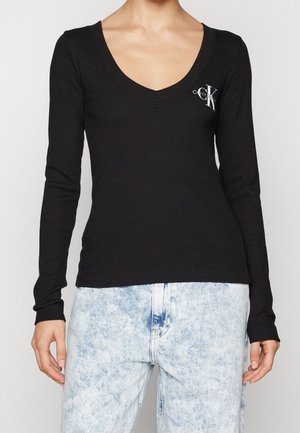 Woman wearing a black long-sleeve V-neck top with a small white CK logo on the chest and light blue acid-wash jeans.