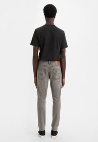 Grey denim jeans with a slim fit, featuring a leather back patch, back pockets, and black shirt, standing on a plain background.