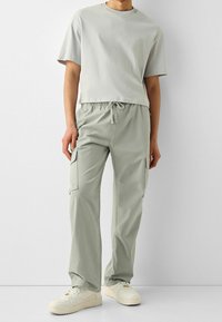 Hellgraues T-Shirt mit kurzen Ärmeln, kombiniert mit hellgrauen Cargohosen mit Seitentaschen und elastischem Bund; dazu weiße Turnschuhe getragen.