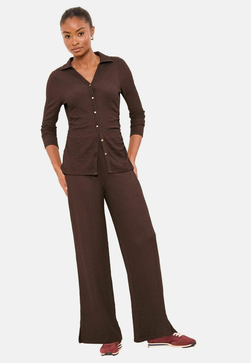 Barna texturált kétdarabos öltözék, amely egy gombos, hosszú ujjú ingből és bő szárú nadrágból áll. Arany színű gombokkal díszítve.