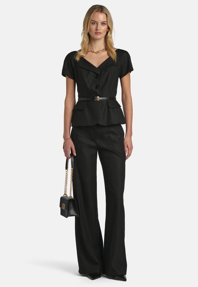 Frau trägt einen schwarzen, maßgeschneiderten Blazer mit kurzen Ärmeln und Gürtel, passende weit geschnittene Hosen, goldenen Schmuck und hält eine schwarze Handtasche mit goldener Kette.