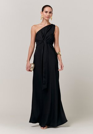 Frau trägt ein einseitig schulterfreies schwarzes Abendkleid mit vorderer Schleife, goldene Ohrringe, Armband, Clutch und Sandalen vor einfachem Hintergrund.