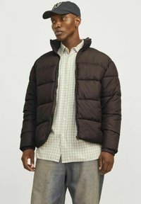 Chaqueta negra acolchada con cuello alto, cierre de cremallera y puños elásticos, llevada sobre una camisa de cuadros y jeans de lavado claro.