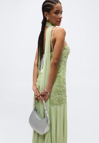 Moteris su pintais plaukais, vilkinti šviesiai žalią gėlių nėriniuota suknelę, laiko mažą pilką rankinę už nugaros, žiūrėdama į šoną.