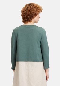 Cardigan corto color teal con maniche lunghe, scollatura rotonda e polsini a volant; realizzato in materiale morbido con una texture liscia.