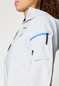 Vaalea harmaa takki, jossa siniset vetoketjuyksityiskohdat ja mustat metalliosat. Takissa on korkea kaulus, näkyvät taskut ja tyylikäs, virtaviivainen muotoilu.