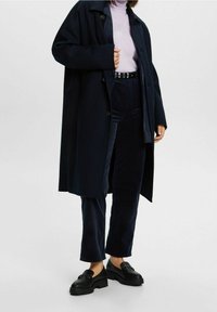 Donkerblauwe lange jas over een lichtpaarse coltrui, gecombineerd met donkerblauwe corduroy broek en zwarte platform loafers. Heeft grote knopen en zakken.